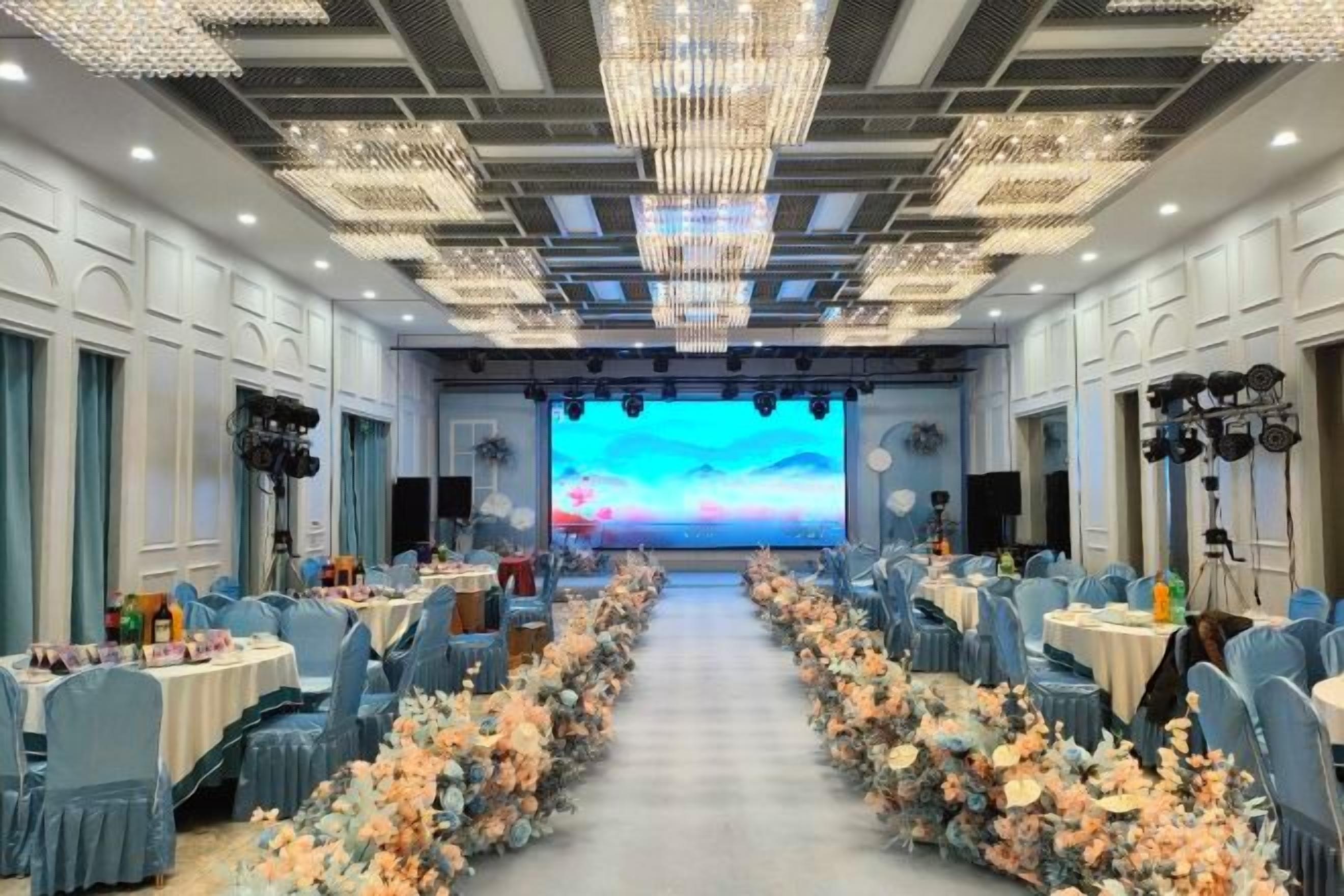 A banquet hall with rows of tables, a stage with a large screen, and a chandelier.