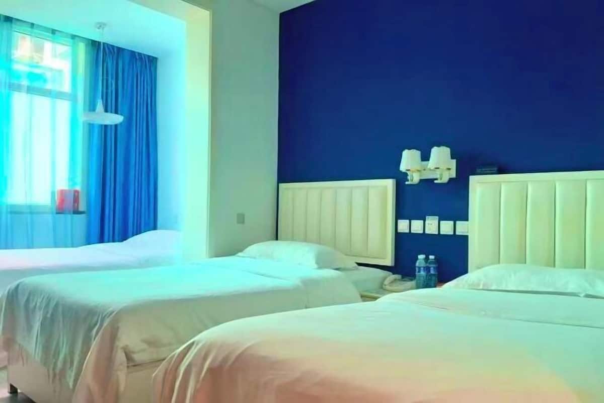 A hotel room with two beds, a window with blue curtains, a white headboard, and a wall-mounted light fixture.
