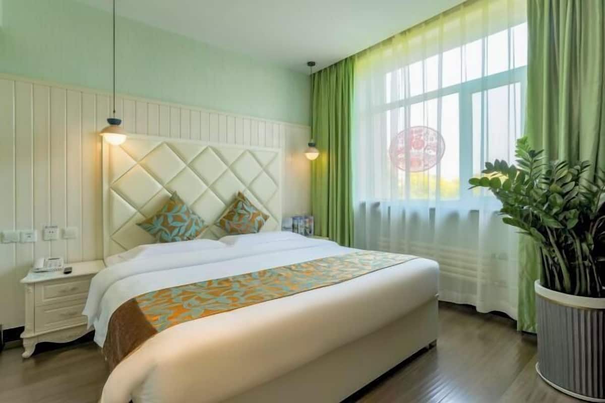 A hotel room with a bed, a nightstand, a potted plant, and a window with curtains.