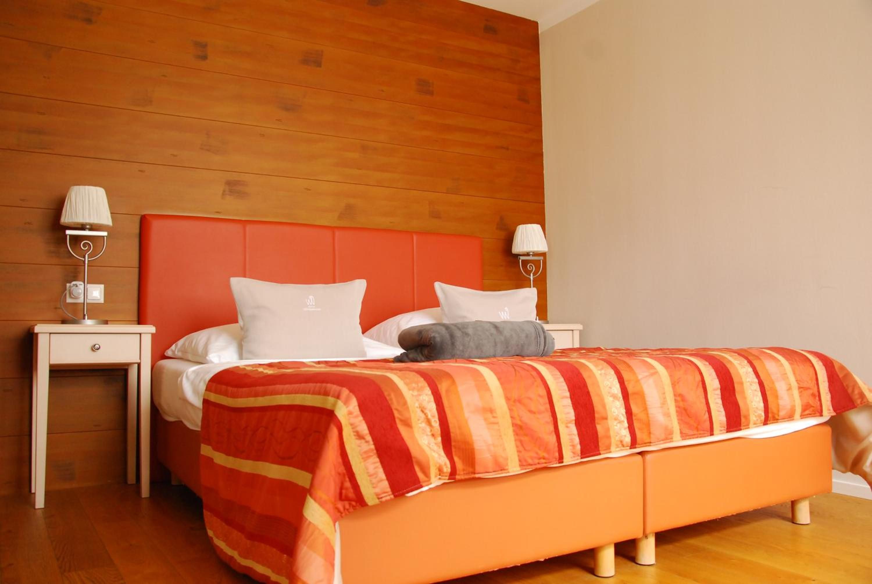 Ein Hotelzimmer mit Holz-Kopfteil, einem Bett mit weißer und orangefarbener Bettwäsche, zwei Nachttischen mit Lampen und einem Holzboden.