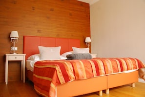 Quarto de hotel com cabeceira de madeira, cama com roupa de cama branca e laranja, duas mesas de cabeceira com abajures e chão de madeira.