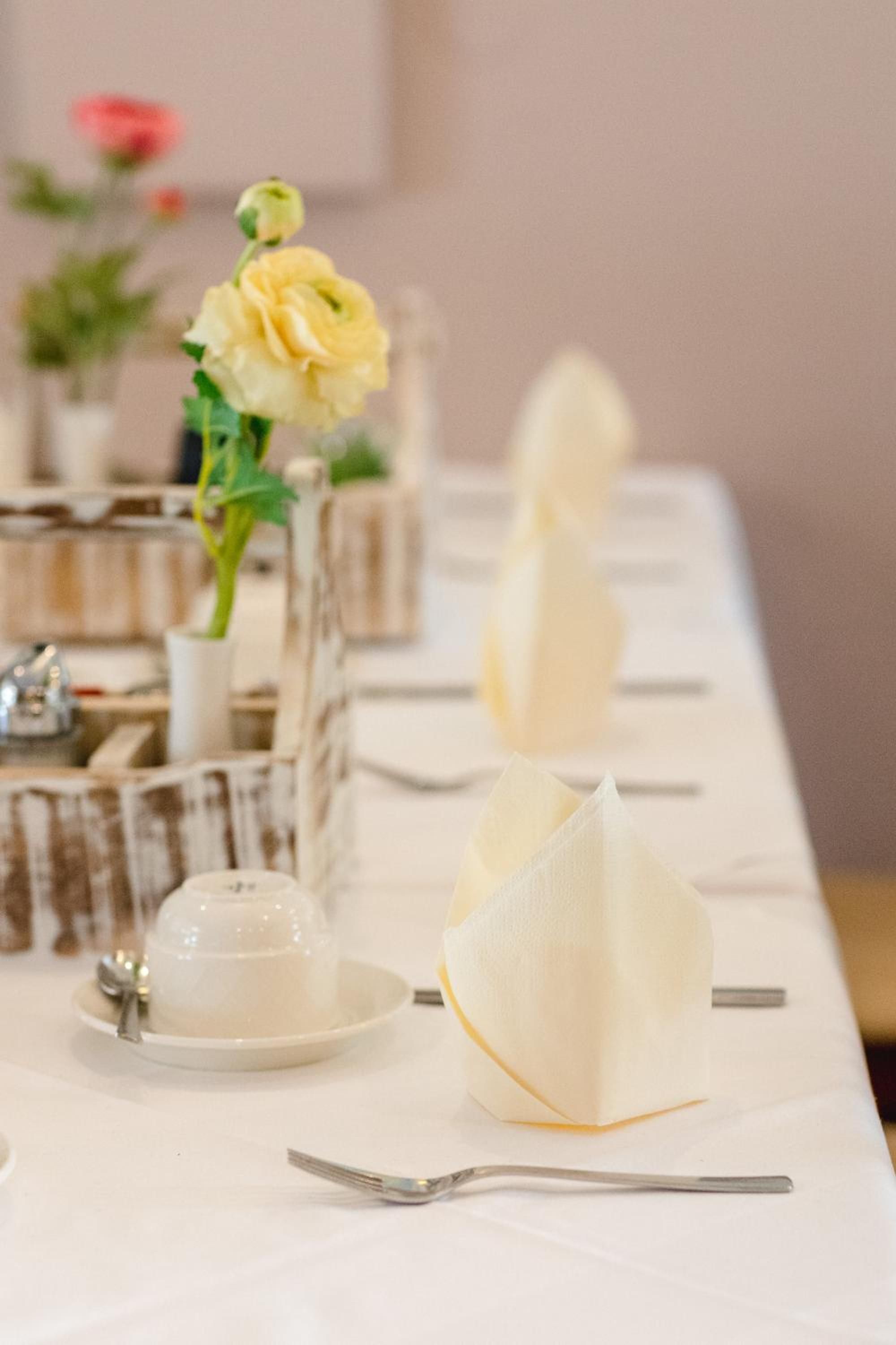 A table set with white napkins, a clear glass vase with a yellow flower, and a silver spoon.