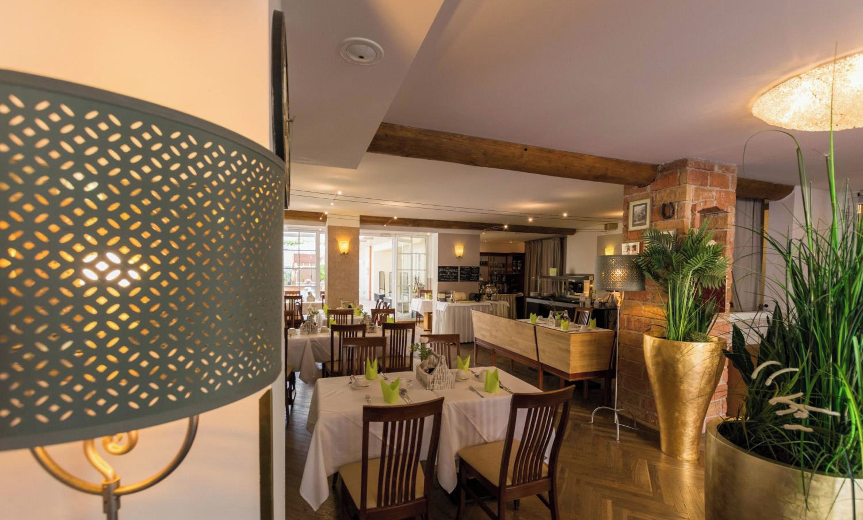 A dining area with tables set for guests, a kitchen in the background, and a large potted plant.