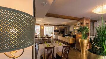 A dining area with tables set for guests, a kitchen in the background, and a large potted plant.