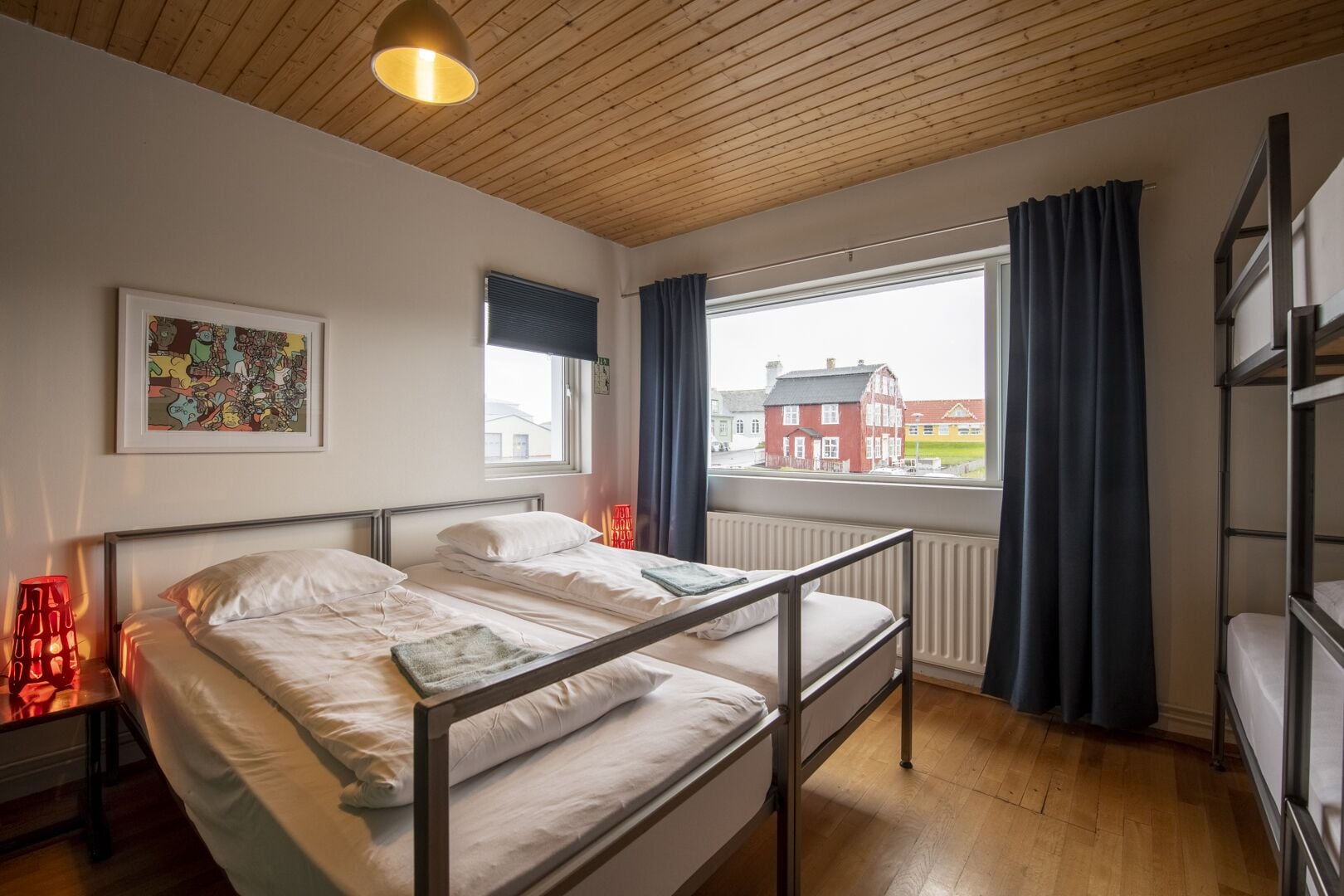 A room with a bunk bed, a single bed, a window with curtains, a radiator, and a framed picture on the wall.