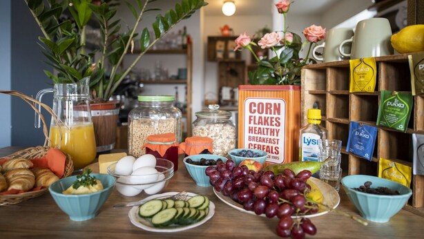 Una mesa de desayuno con diferentes alimentos, una jarra de jugo de naranja y una caja de copos de maíz.