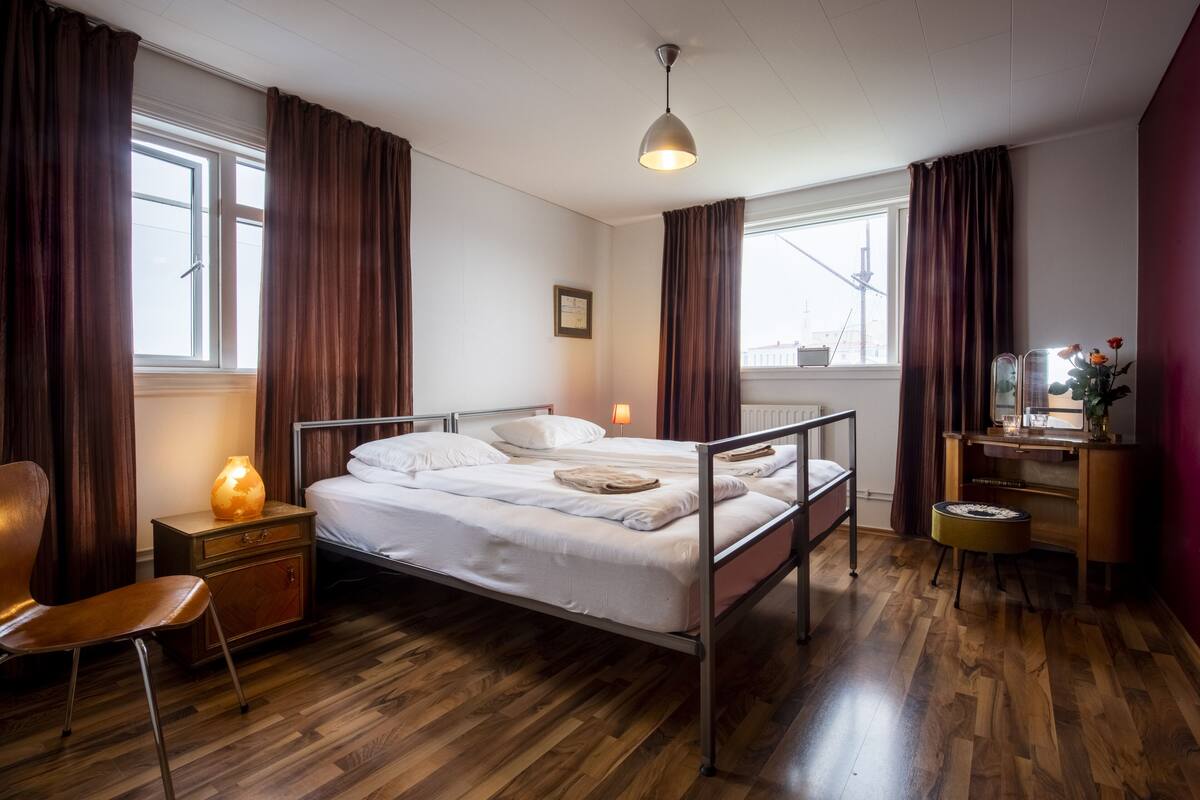 A bedroom with a metal bed frame, wooden flooring, a bedside table with a lamp, a chair, and a dresser.