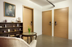 Un vestíbulo de hotel con un mostrador de recepción de madera, una mesa de vidrio con un jarrón de flores, una estantería con diversos objetos y dos puertas de madera.
