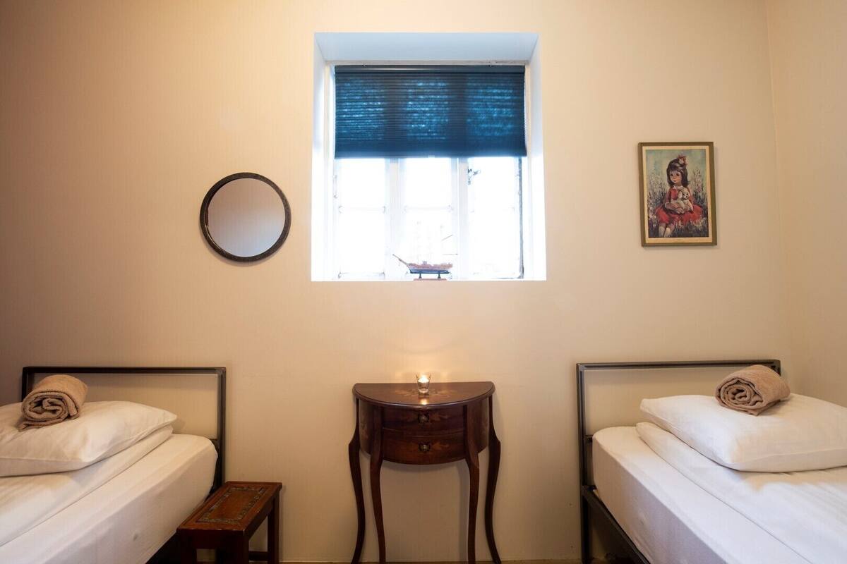 A hotel room with two single beds, a small wooden table, a round mirror, a window with a dark blind, and a framed picture on the wall.