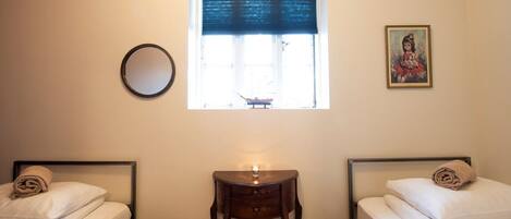 A hotel room with two single beds, a small wooden table, a round mirror, a window with a dark blind, and a framed picture on the wall.