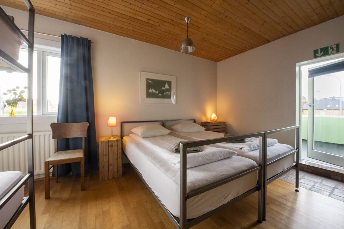 A bedroom with a bunk bed, a chair, bedside lamps, a window with curtains, and a framed picture on the wall.