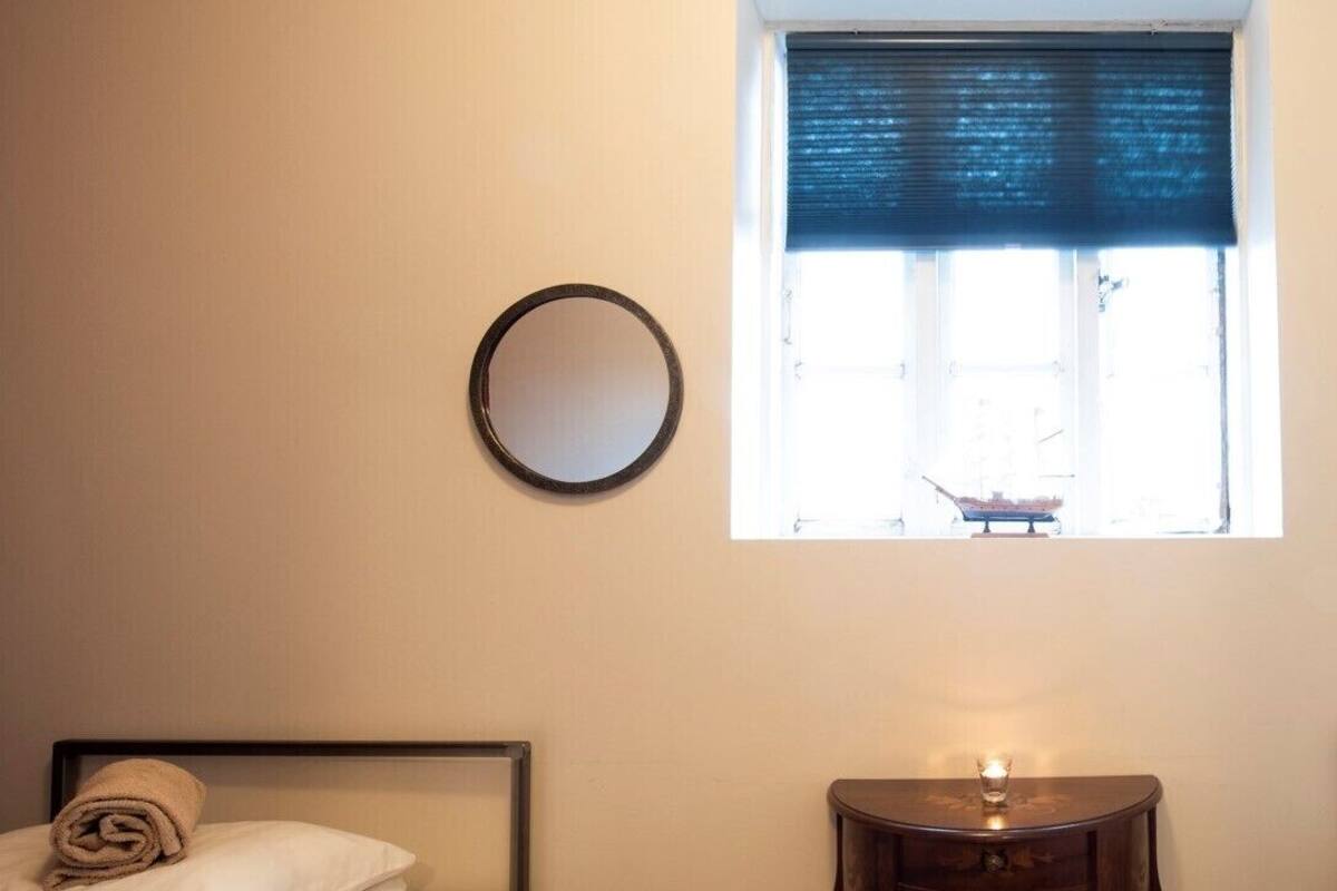 A bedroom with a bed, a bedside table with a lamp, a window with a view, and a round mirror on the wall.