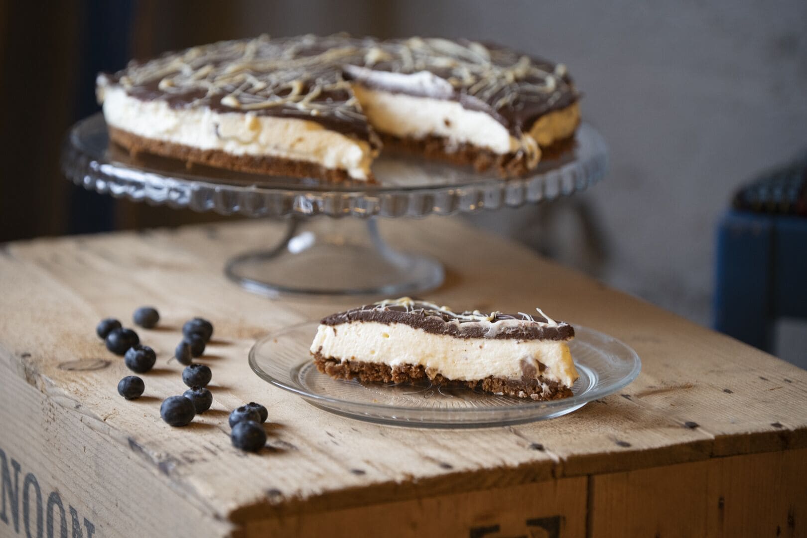 A cheesecake on a glass stand with a slice on a separate plate, blueberries on a wooden surface, and a blurred background.