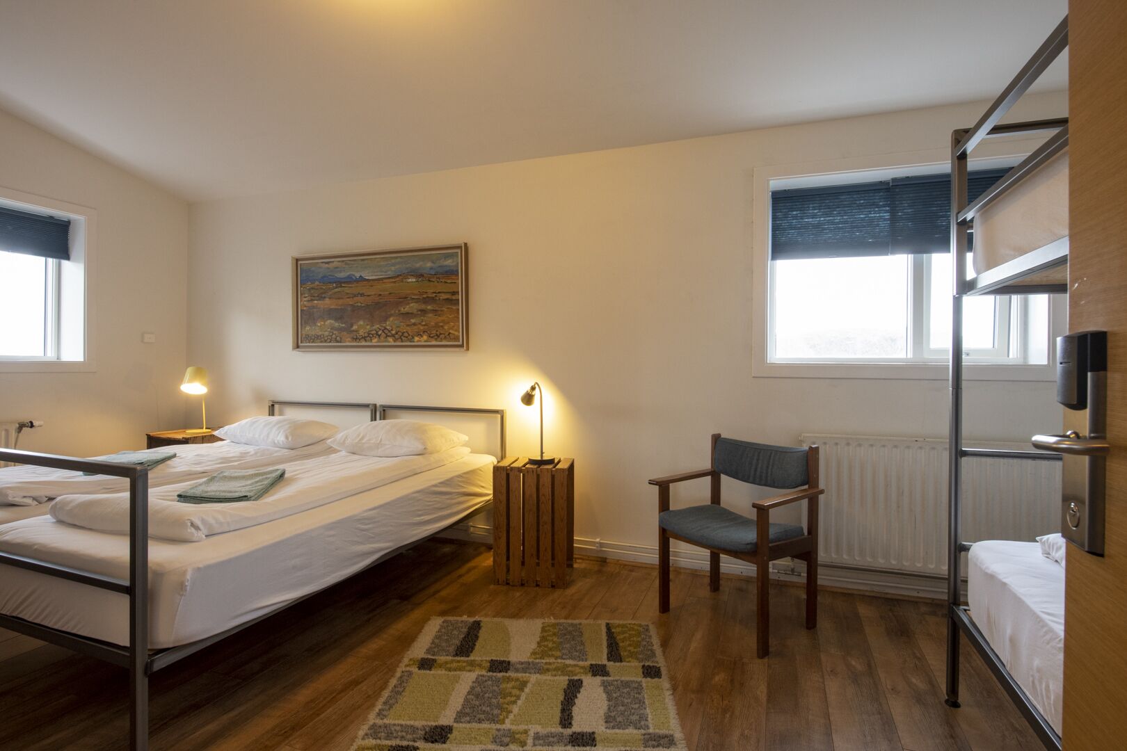 A hotel room with two beds, a chair, a painting, and a radiator.