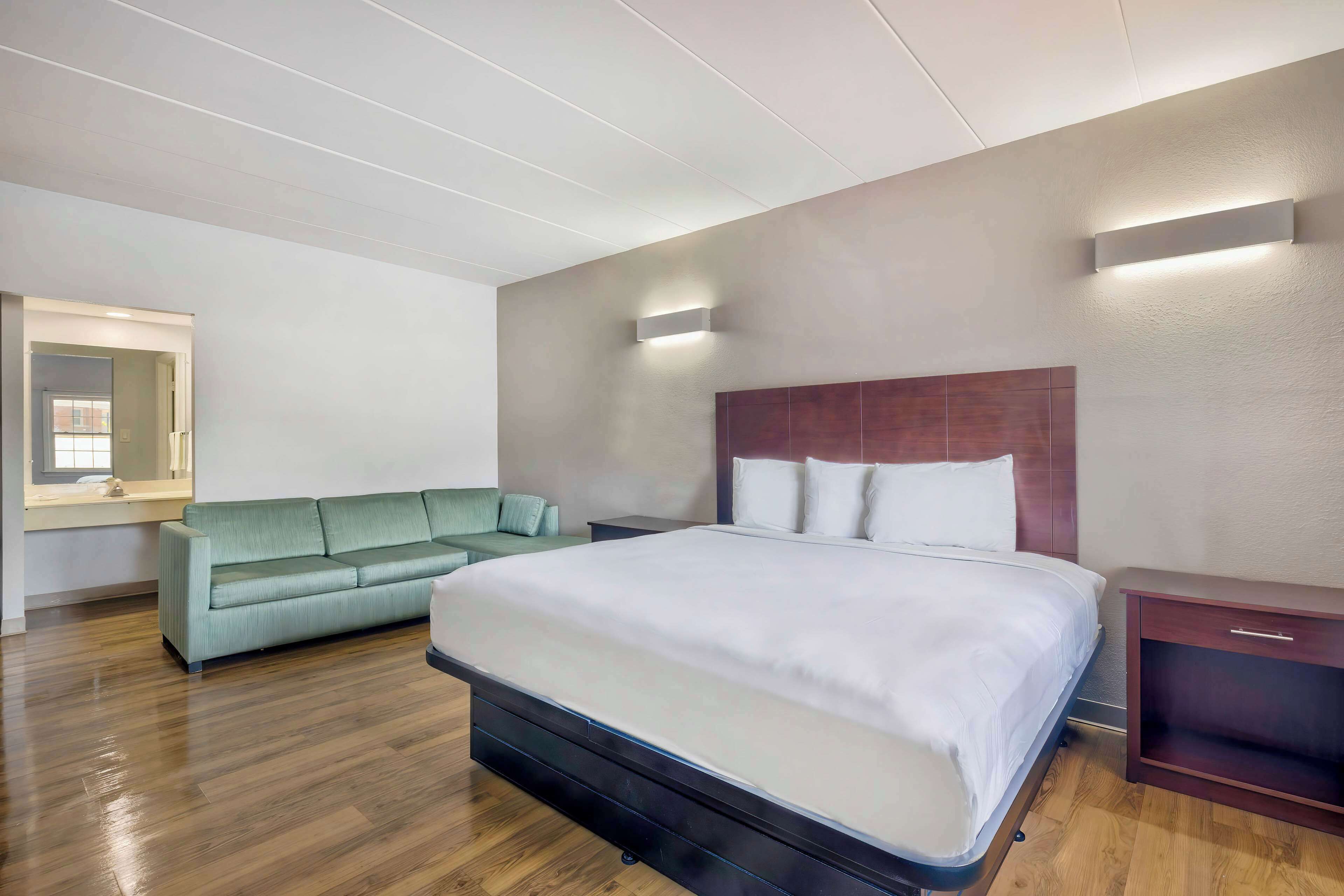 Habitación de hotel moderna con una cama grande, un sofá verde, piso de madera e iluminación integrada.
