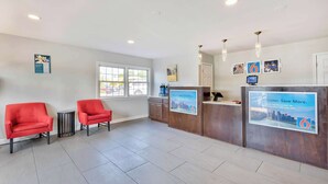 A reception area with two red chairs, a small round table, a wooden counter with a digital screen, and a large poster advertising a mobile app. - Motel 6 Falls Church, VA - Arlington Boulevard (Falls Church)