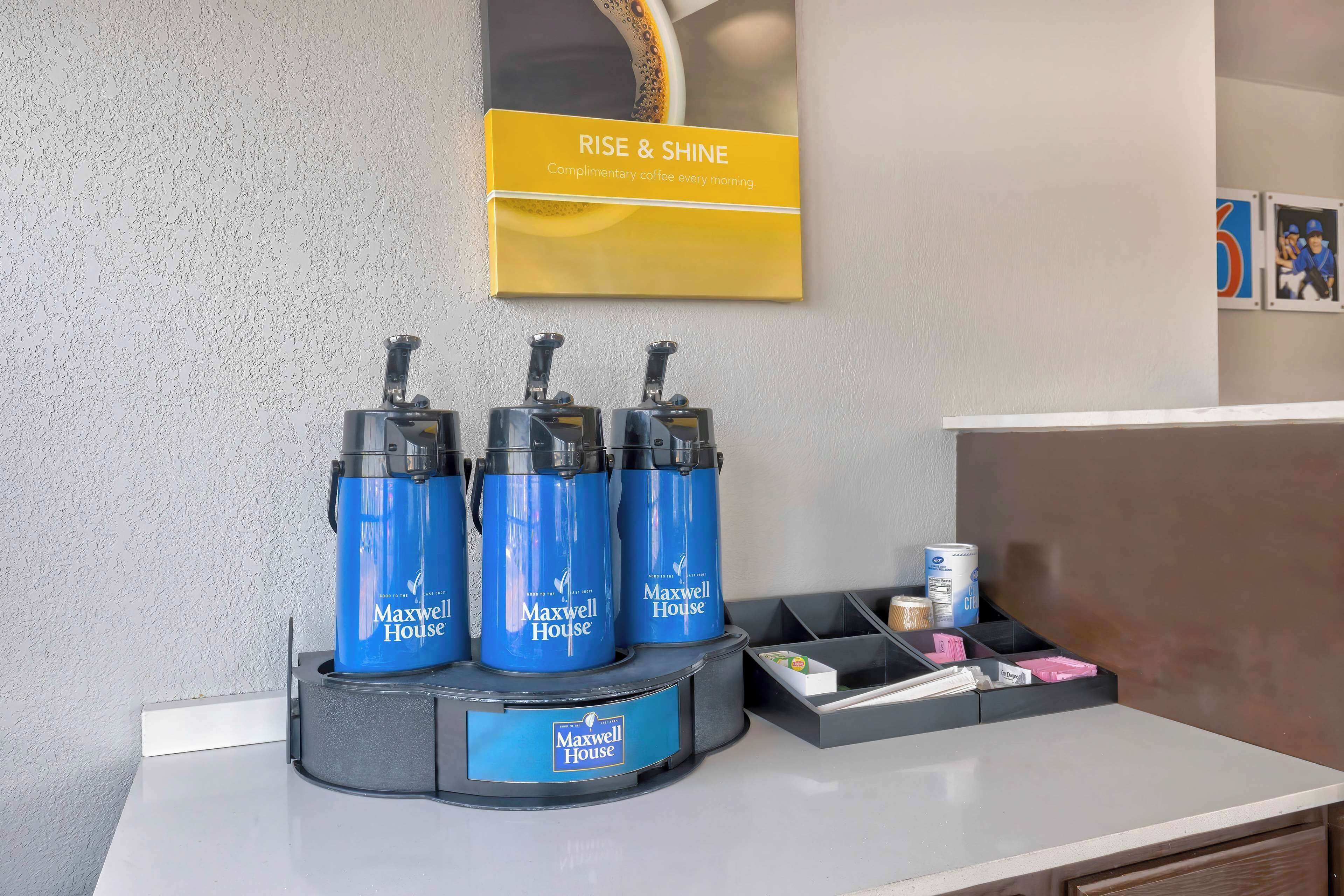 A coffee station with three blue Maxwell House coffee makers on a gray stand, a yellow sign above reading "RISE & SHINE," and a small tray with coffee cups and a can of creamer.