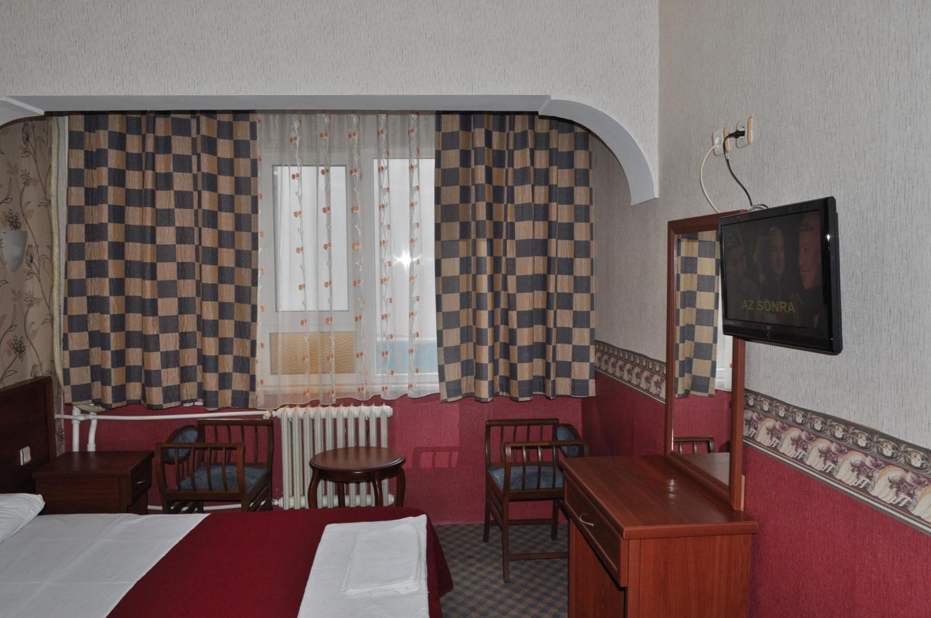 Hotelkamer met gordijn met ruitpatroon, een bed, bureau, twee stoelen, radiator en een televisie aan de muur.