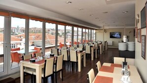 Un restaurante con varias mesas y sillas, amplias ventanas que dan a una vista de la ciudad y un televisor en la pared.