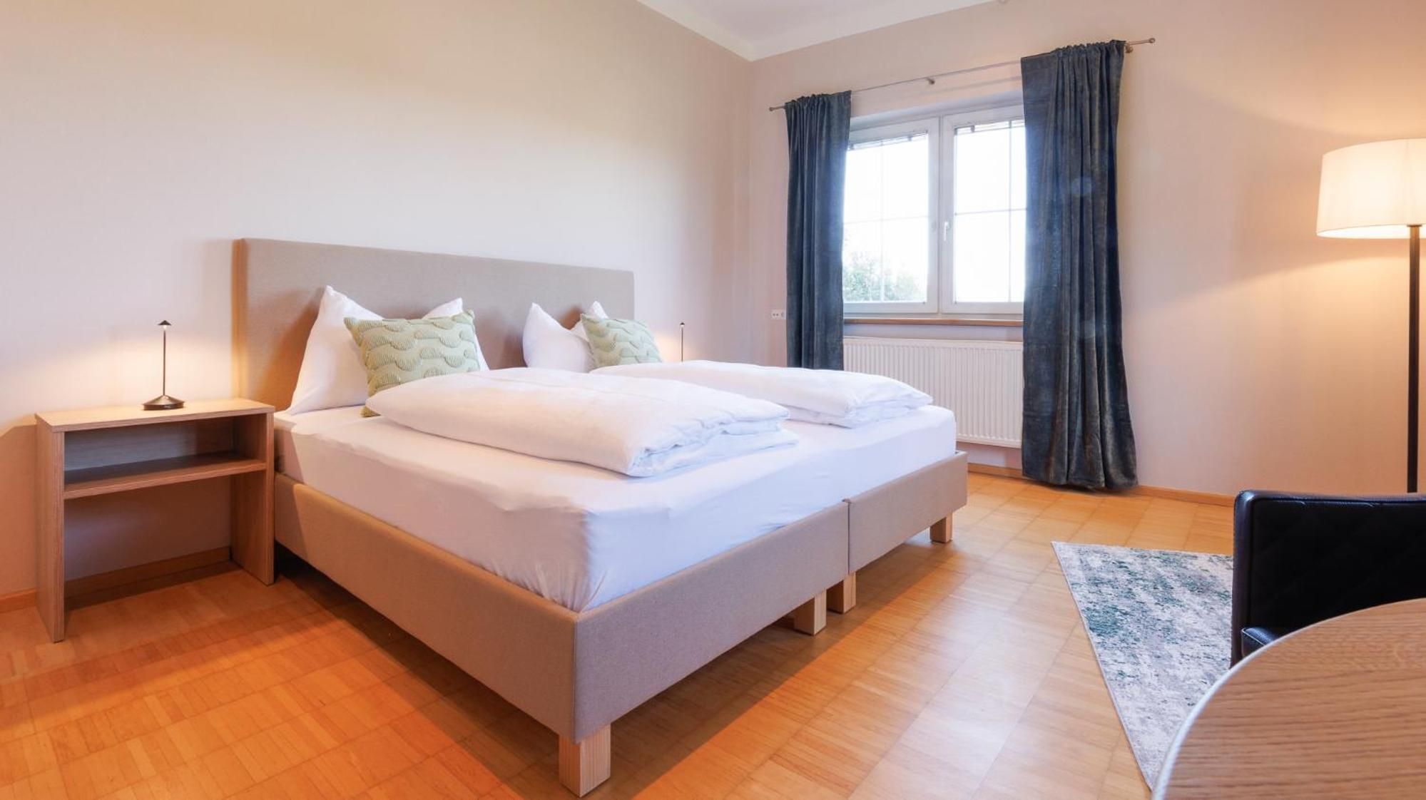 Une chambre d’hôtel avec un lit, des tables de chevet, une lampe, une chaise et une fenêtre avec des rideaux.