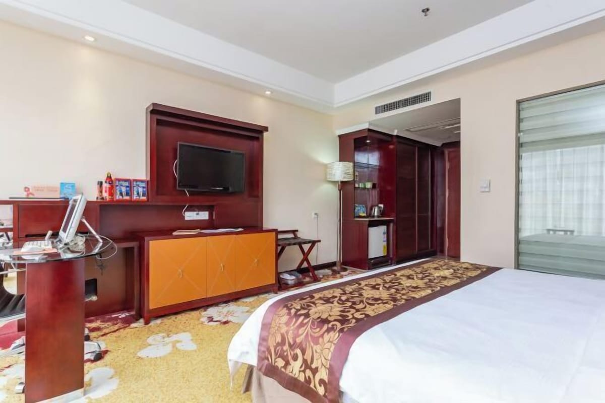 A hotel room with a bed, a television mounted on the wall, a desk with a lamp, and a wardrobe.