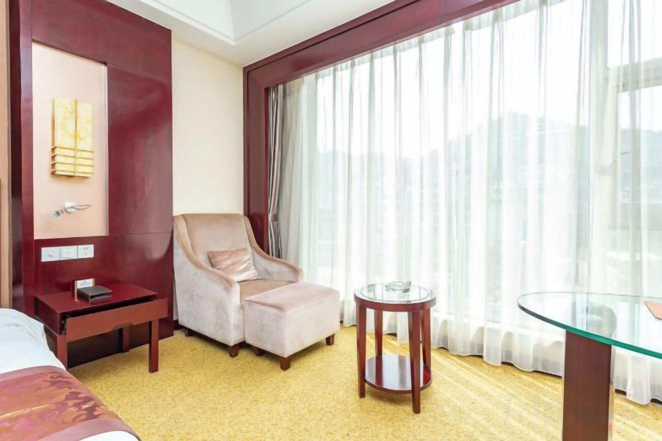 A hotel room with a bed, a chair, a small table, and a large window with curtains.