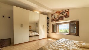 A bedroom with a large wardrobe, a bed with a patterned duvet, a window with a view, and artwork on the wall. - Villa Leonardo (Sarteano)