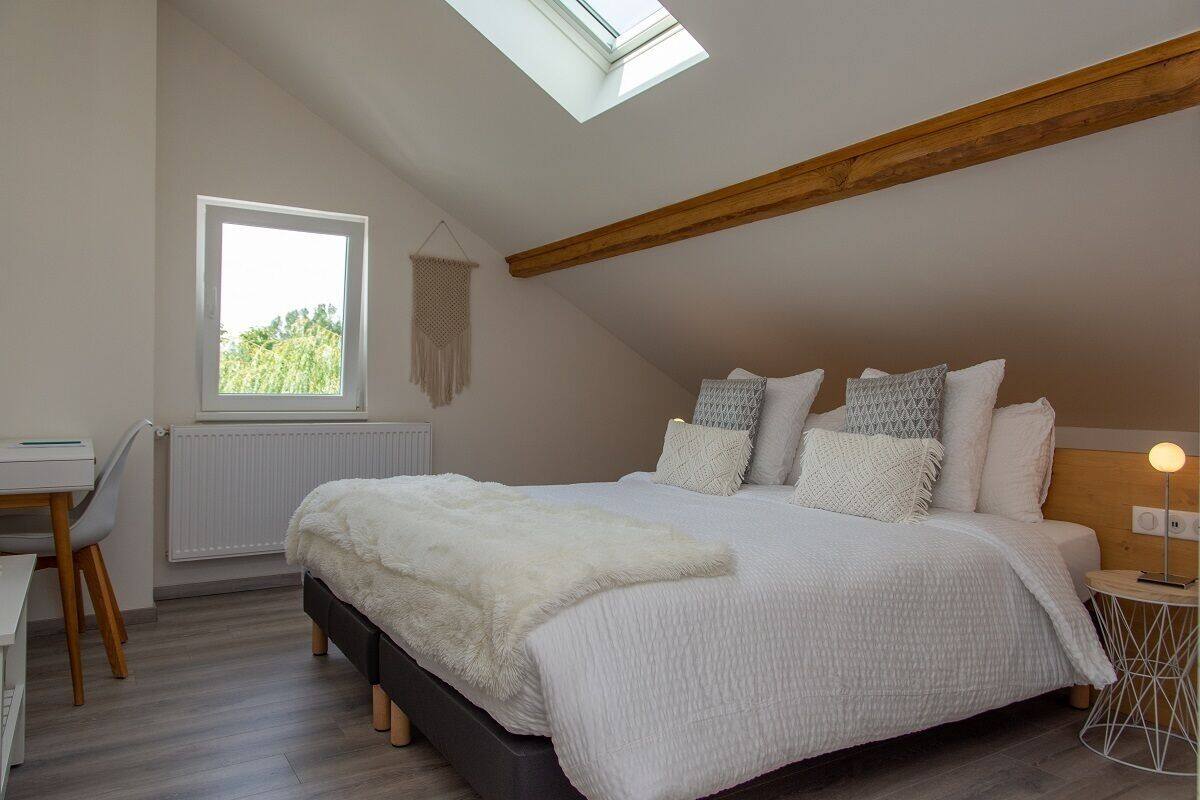A bedroom with a skylight, a bed with multiple pillows, a bedside table with a lamp, and a chair.