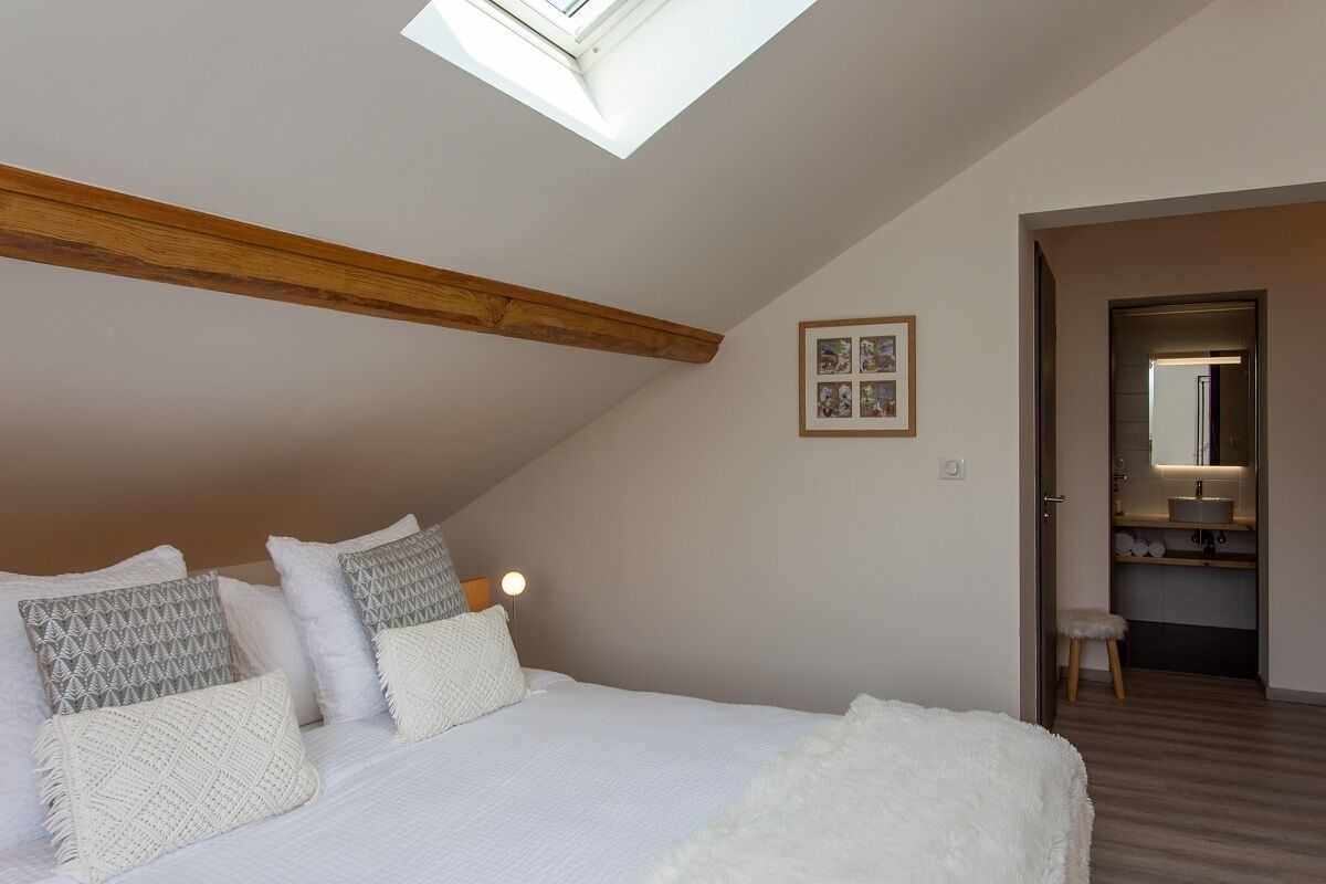 Une chambre avec un plafond en pente, un puits de lumière, un lit avec une literie blanche, et une petite salle de bain visible à travers une porte ouverte.