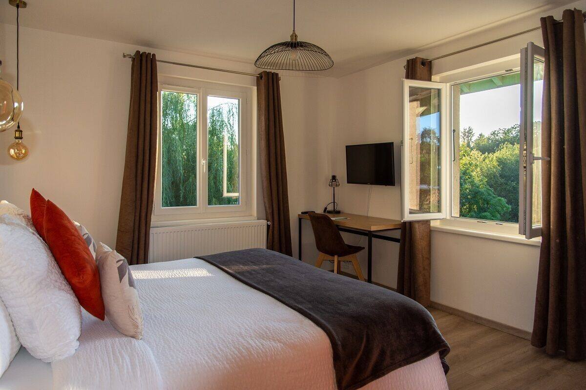 A bedroom with a bed, a desk, a chair, a TV, and large windows with curtains.