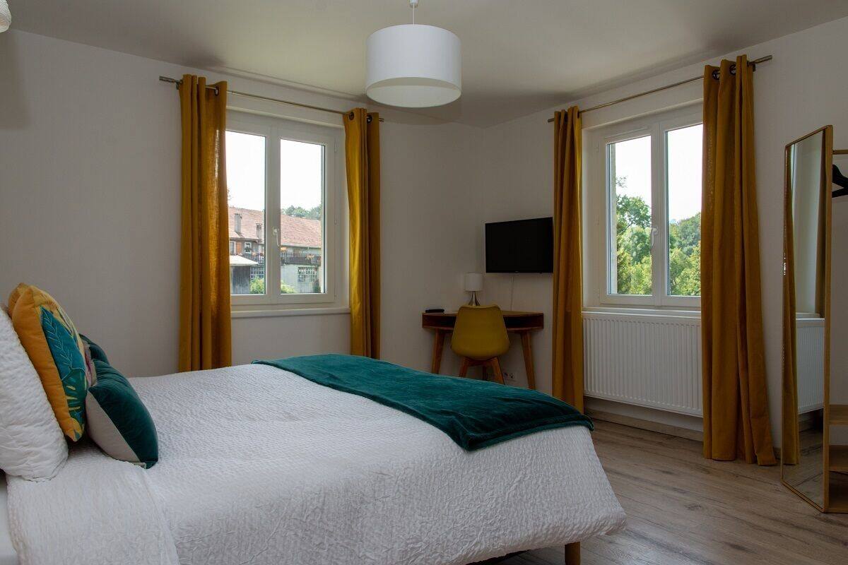 A bedroom with a bed, a desk, a chair, a TV, and large windows with curtains.