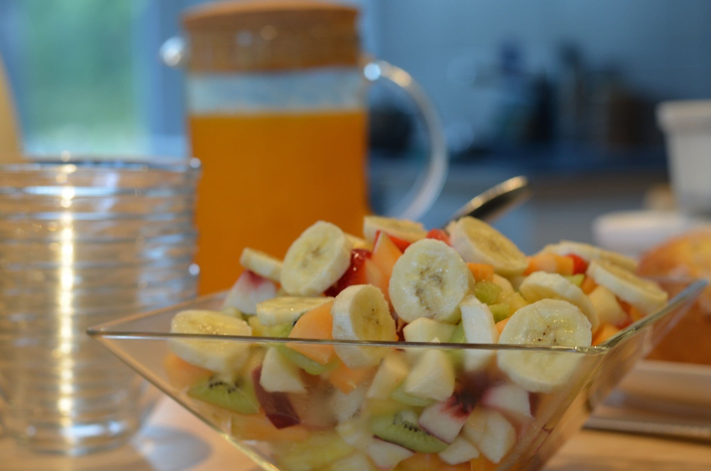 A glass bowl filled with a fruit salad, a glass of orange juice, and a pitcher of orange juice in the background.
