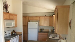 A compact kitchen with wooden cabinets, a white refrigerator, a microwave, an oven, and a toaster. There are wine bottles and a coffee maker on the counter. - The Cozy Cabin LaPine (La Pine)