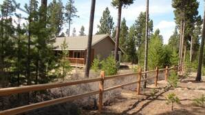 A cabin surrounded by tall pine trees, with a wooden deck and a fenced area. - The Cozy Cabin LaPine (La Pine)