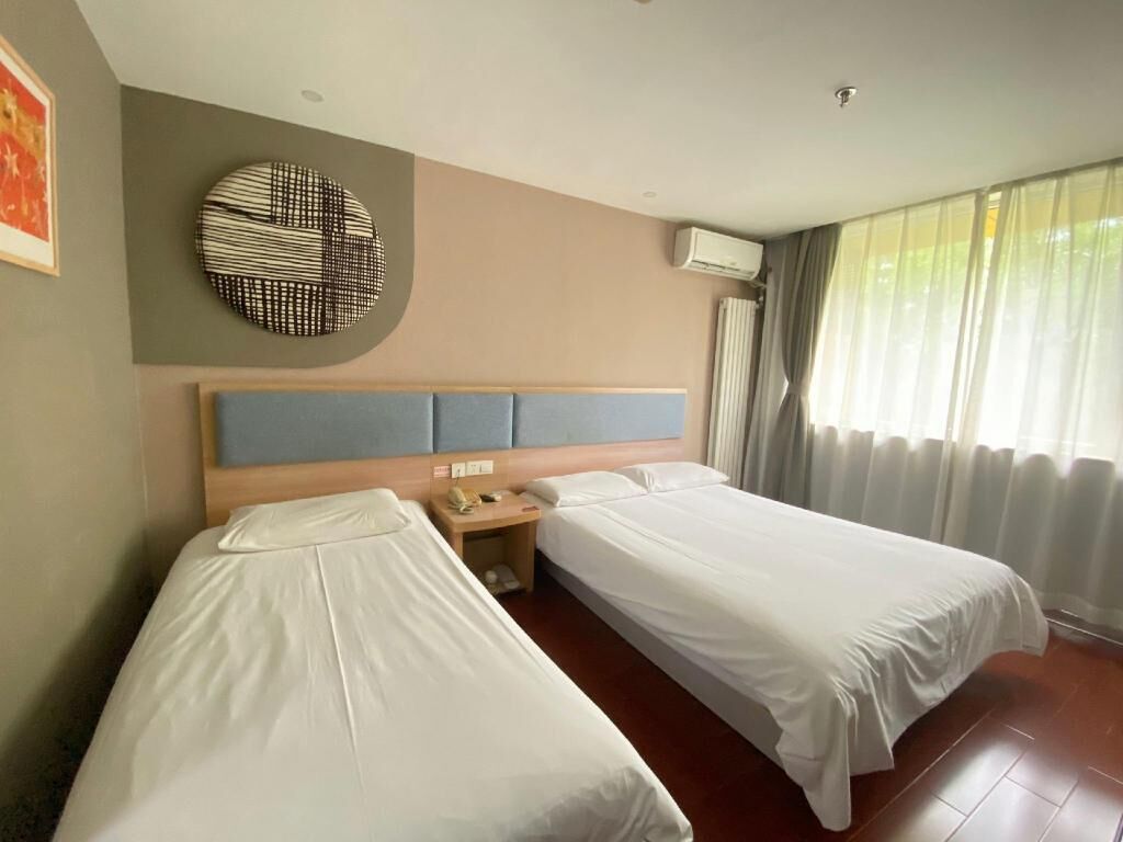 Habitación de hotel con dos camas individuales, un adorno circular en la pared, un equipo de aire acondicionado y una ventana con cortinas.