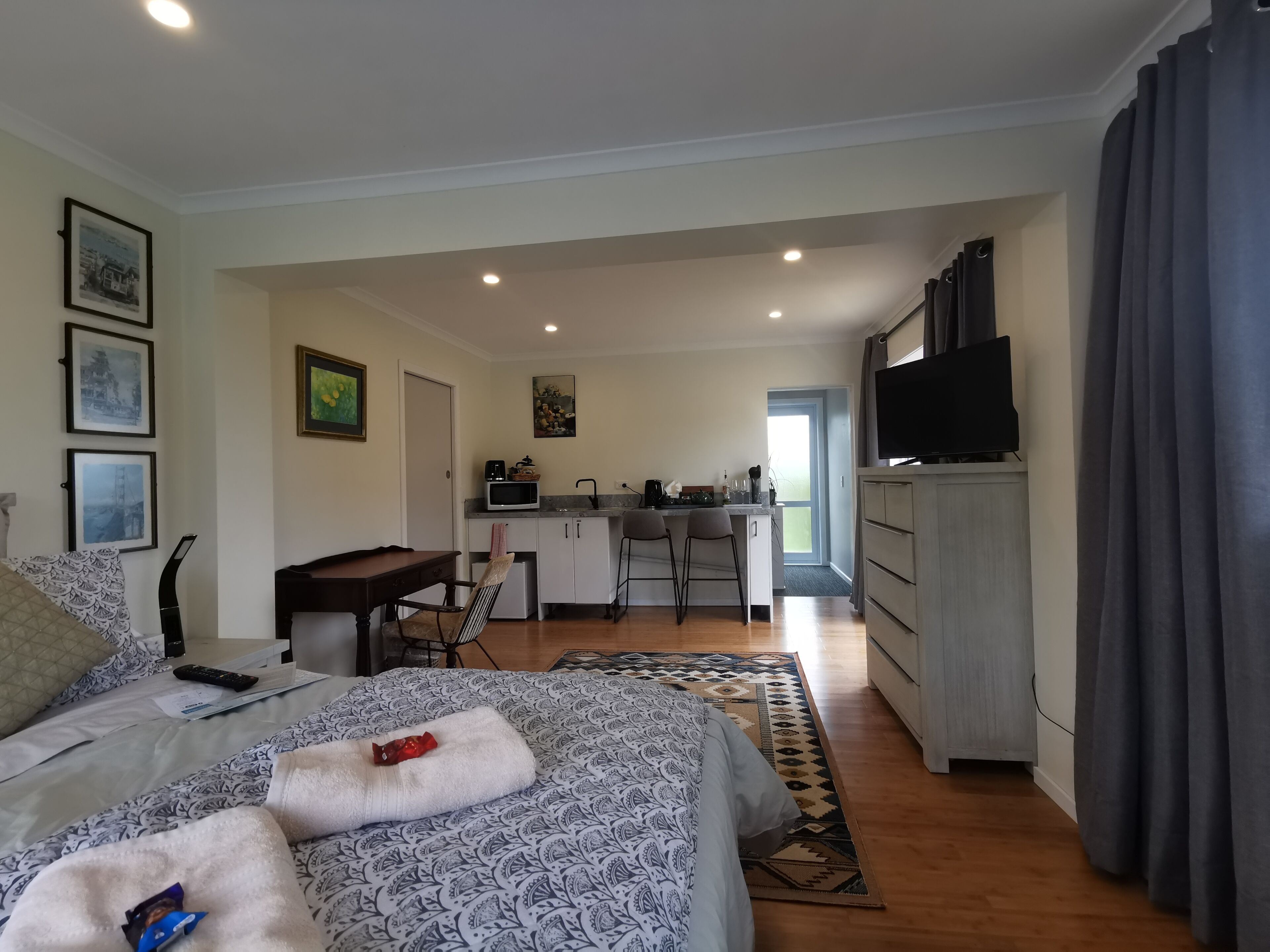 A bedroom with a bed, bedside table, and a desk with a chair. There is a kitchenette with a microwave, coffee maker, and a TV.