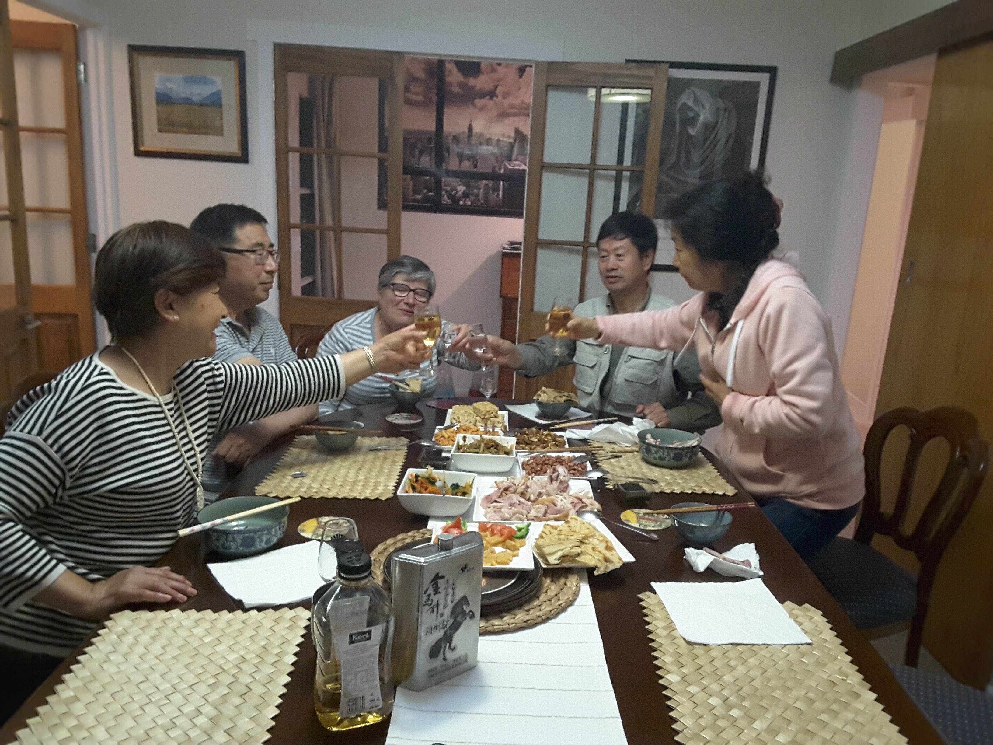 A group of people sitting around a table with various dishes and drinks, in a room with wooden furniture and a view of the outside through a window.