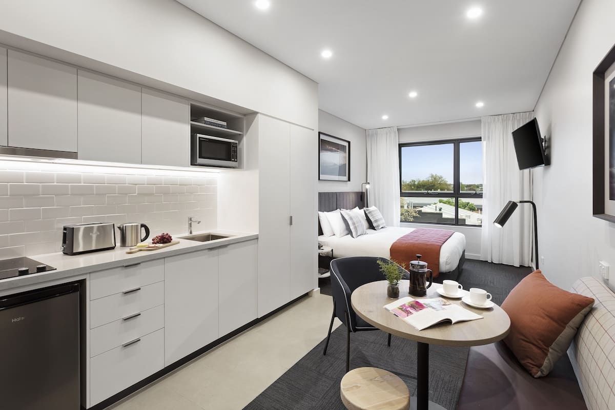A modern hotel room with a kitchenette, a dining area, a bed with a view, and a TV mounted on the wall.