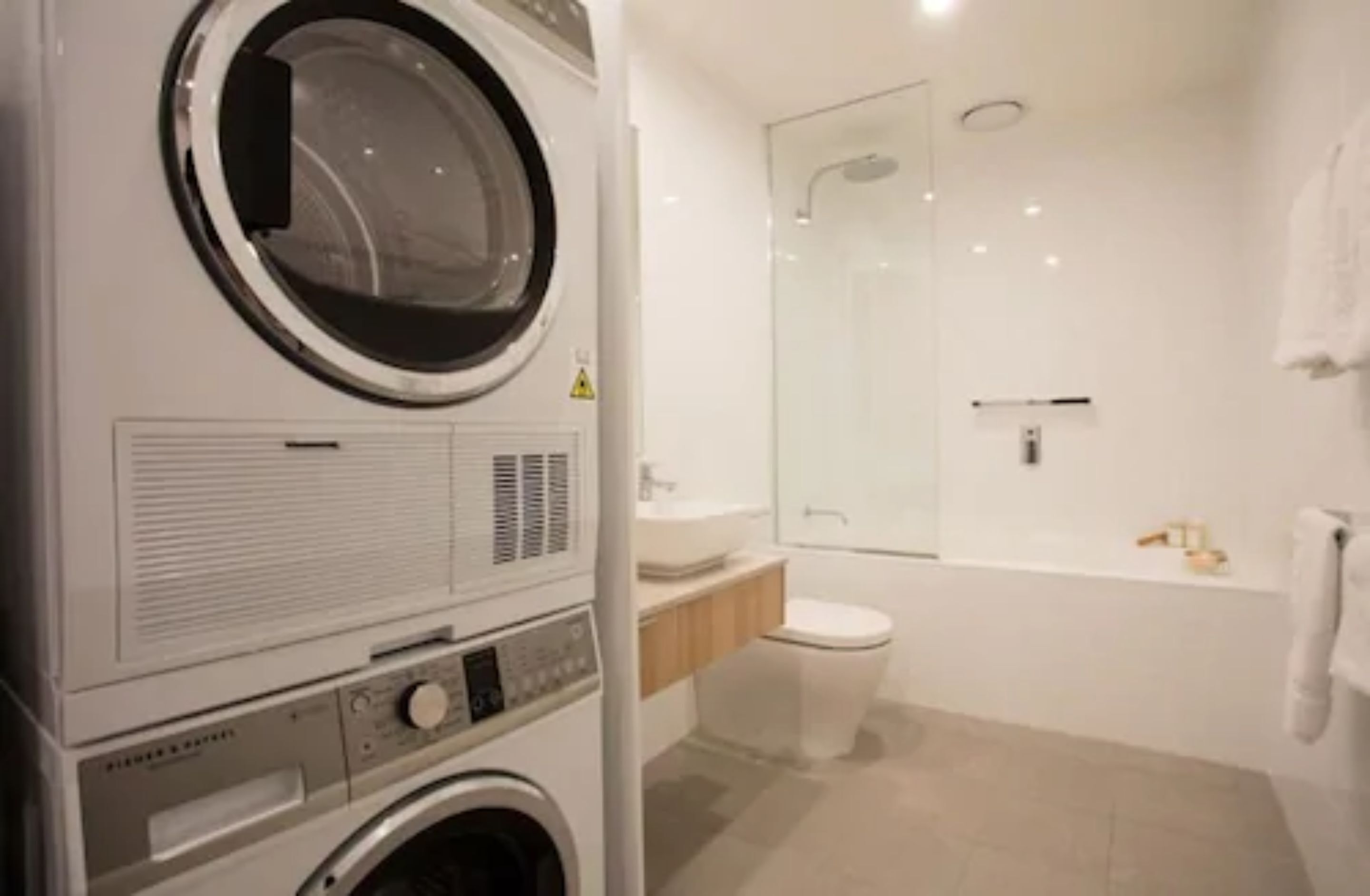 Zona de lavandería con lavadora y secadora de carga frontal, cabina de ducha con mampara de vidrio, inodoro y bañera.