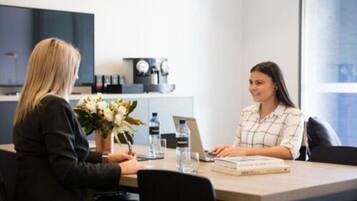 Two people in a meeting room with a table, water bottles, a laptop, and a vase with flowers.
