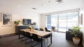 Quarto de hotel moderno com uma janela grande, televisão de tela plana, área de refeições com mesa e cadeiras, e uma planta em vaso.