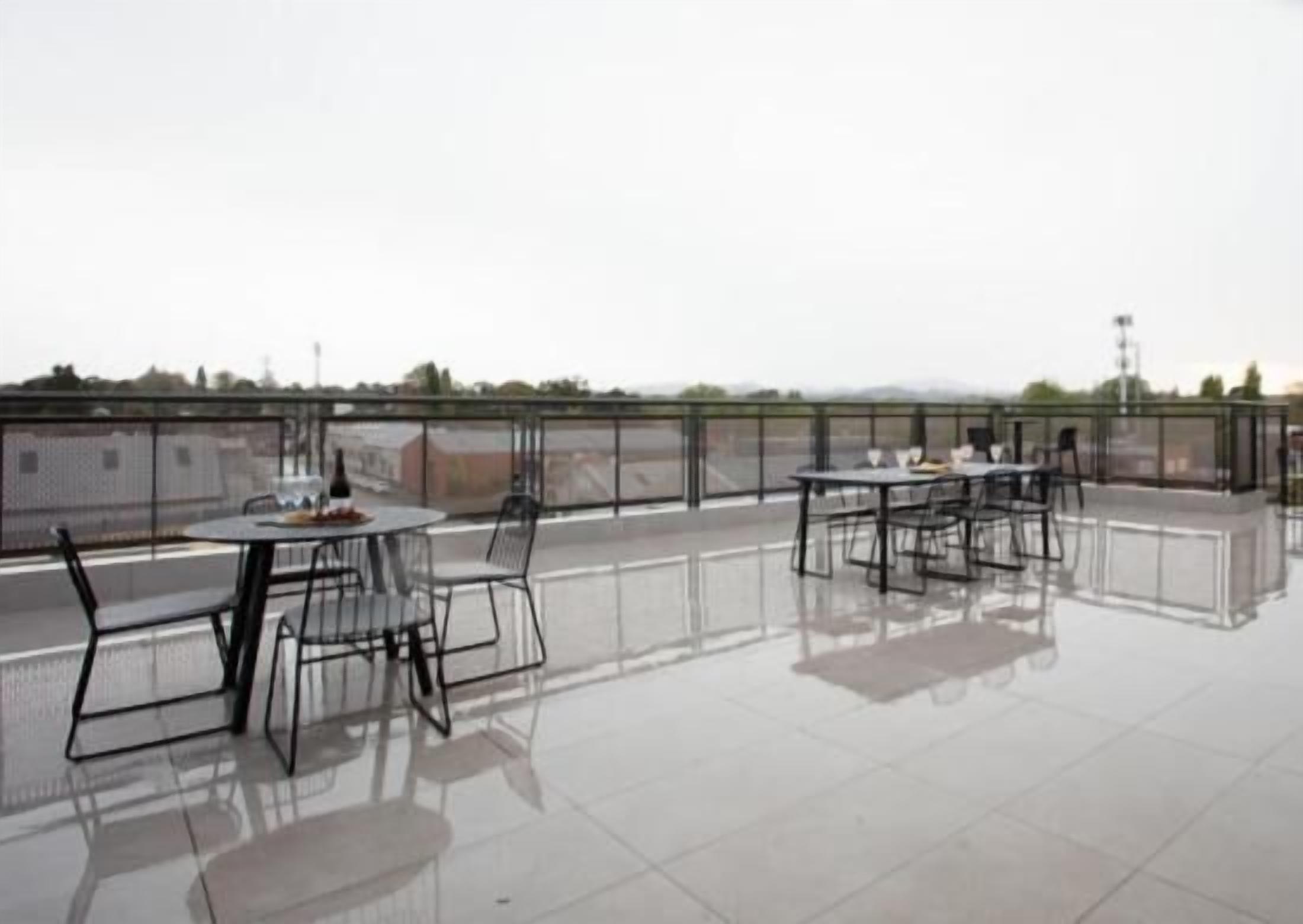 A rooftop terrace with tables and chairs, a glass railing, and a view of the cityscape.