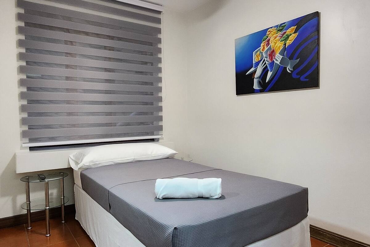 A bedroom with a bed, a ceiling fan, a window with blinds, and a painting on the wall.