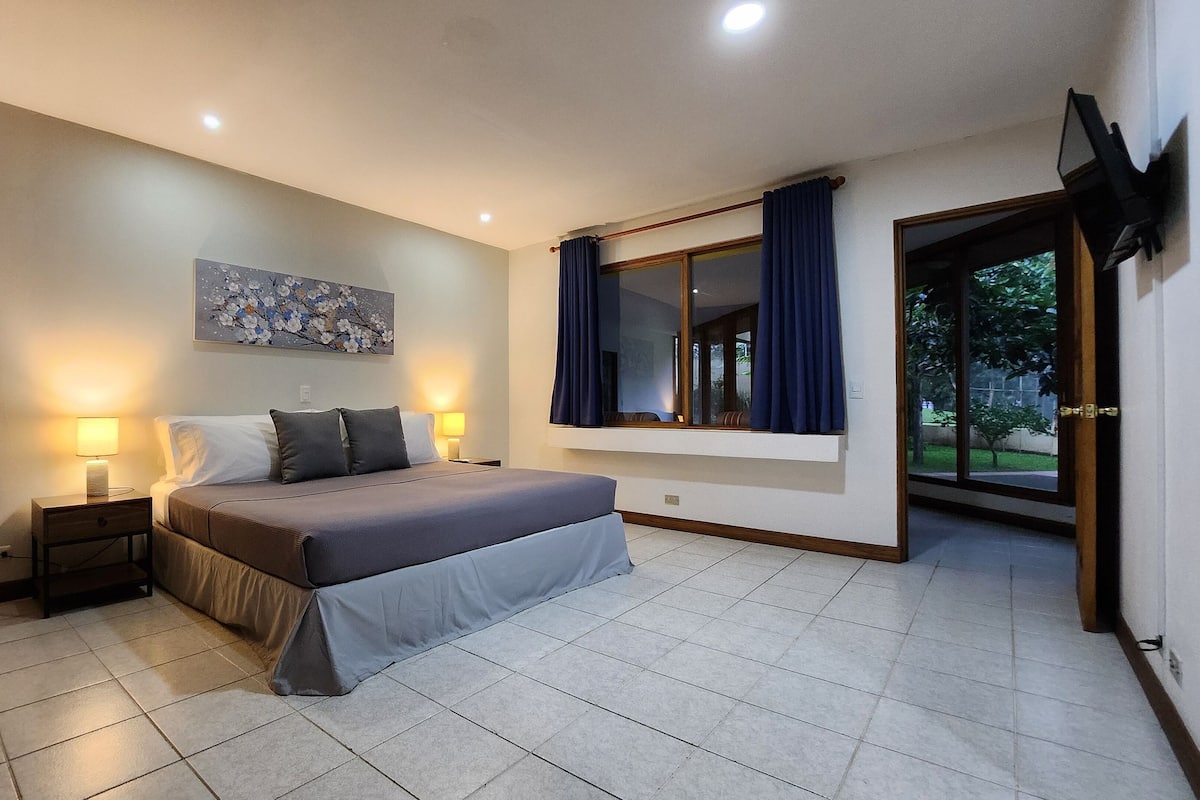 A bedroom with a bed, nightstands, a window with curtains, a painting on the wall, and a door leading outside.