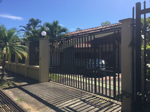 Uma entrada com portão, um veículo no interior, palmeiras e um edifício com telhado em cerâmica.