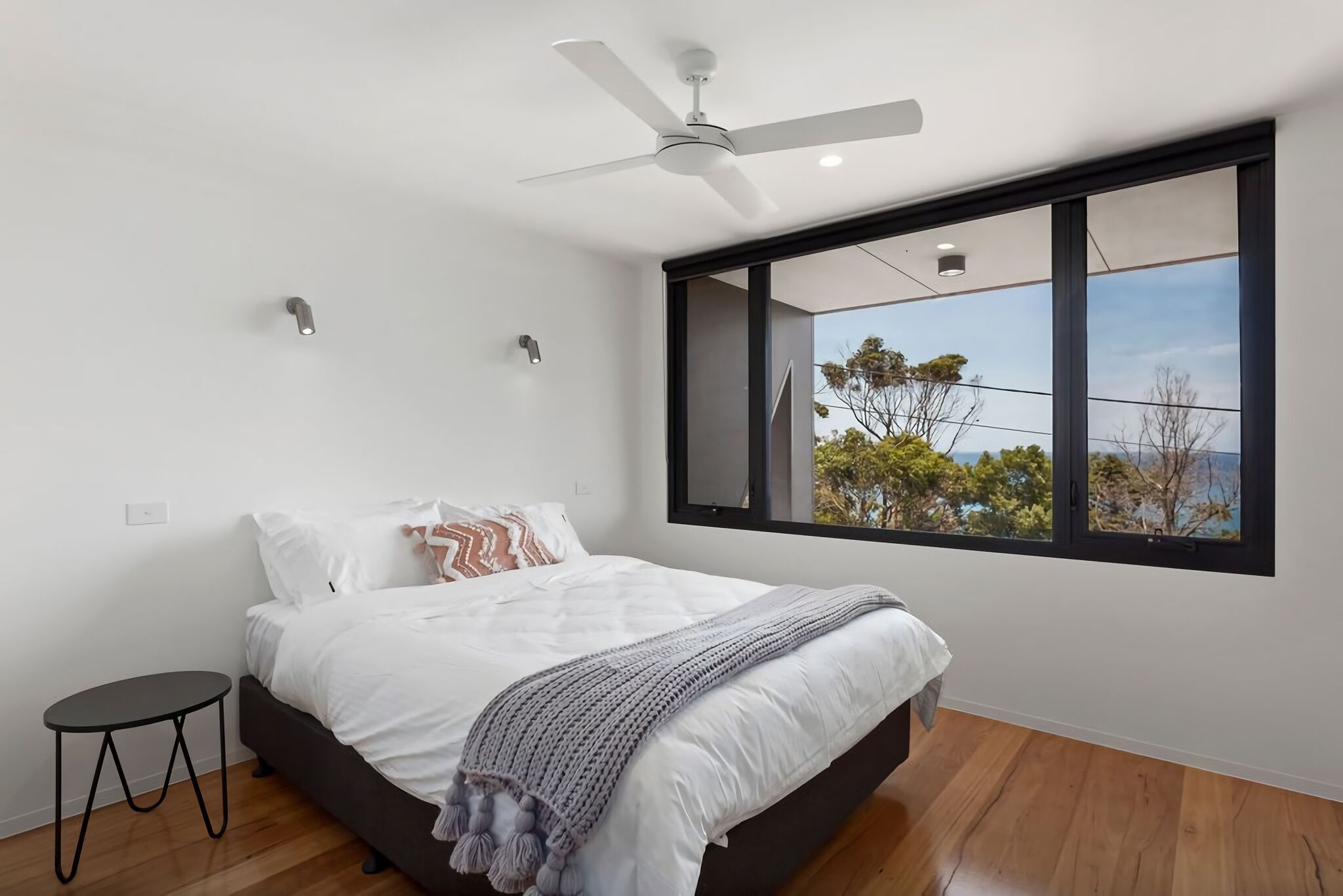 A modern bedroom with a large window, a ceiling fan, and a small bedside table.