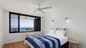 Een slaapkamer met een groot raam, een bed met een blauw-wit geruit dekbed, een plafondventilator en een klein houten krukje.