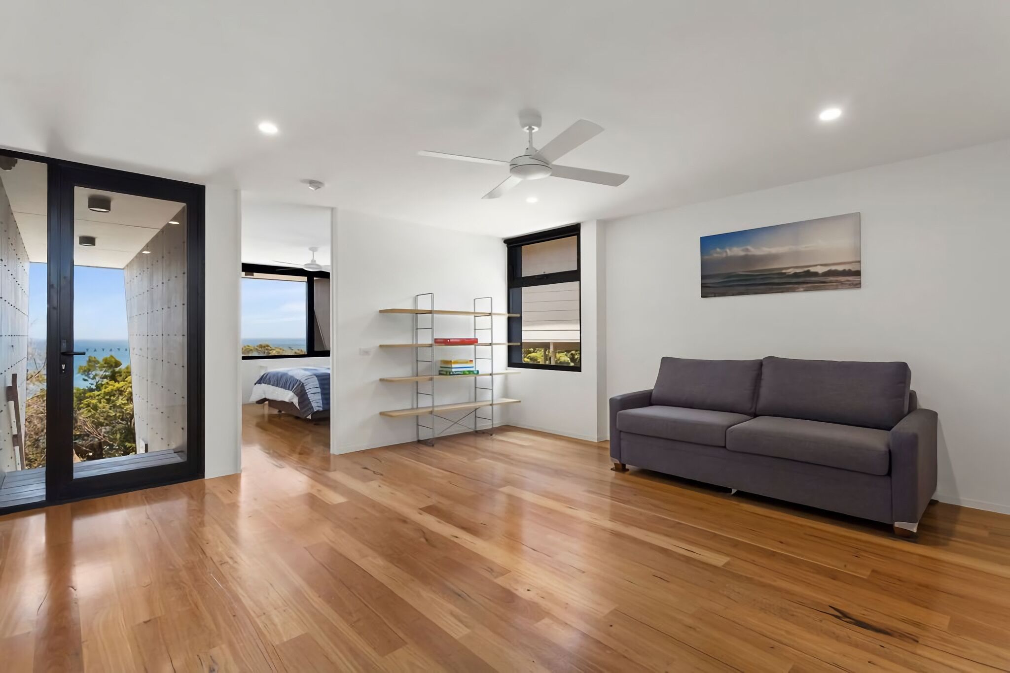 A modern living room with a grey sofa, wooden flooring, a ceiling fan, and a bookshelf.