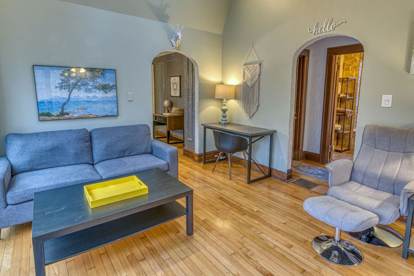 Una sala de estar con un sofá, una mesa de centro, una mesa de comedor y una silla. Hay un cuadro en la pared y una lámpara sobre la mesa.
