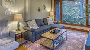A living room with a grey sofa, a coffee table with books, a lamp, and a view of a stone wall and trees through a window. - Secluded, Fenced Cottage, Hot Tub, Fire Pit, Dog Friendly, Nat’l Park (Miller Beach)